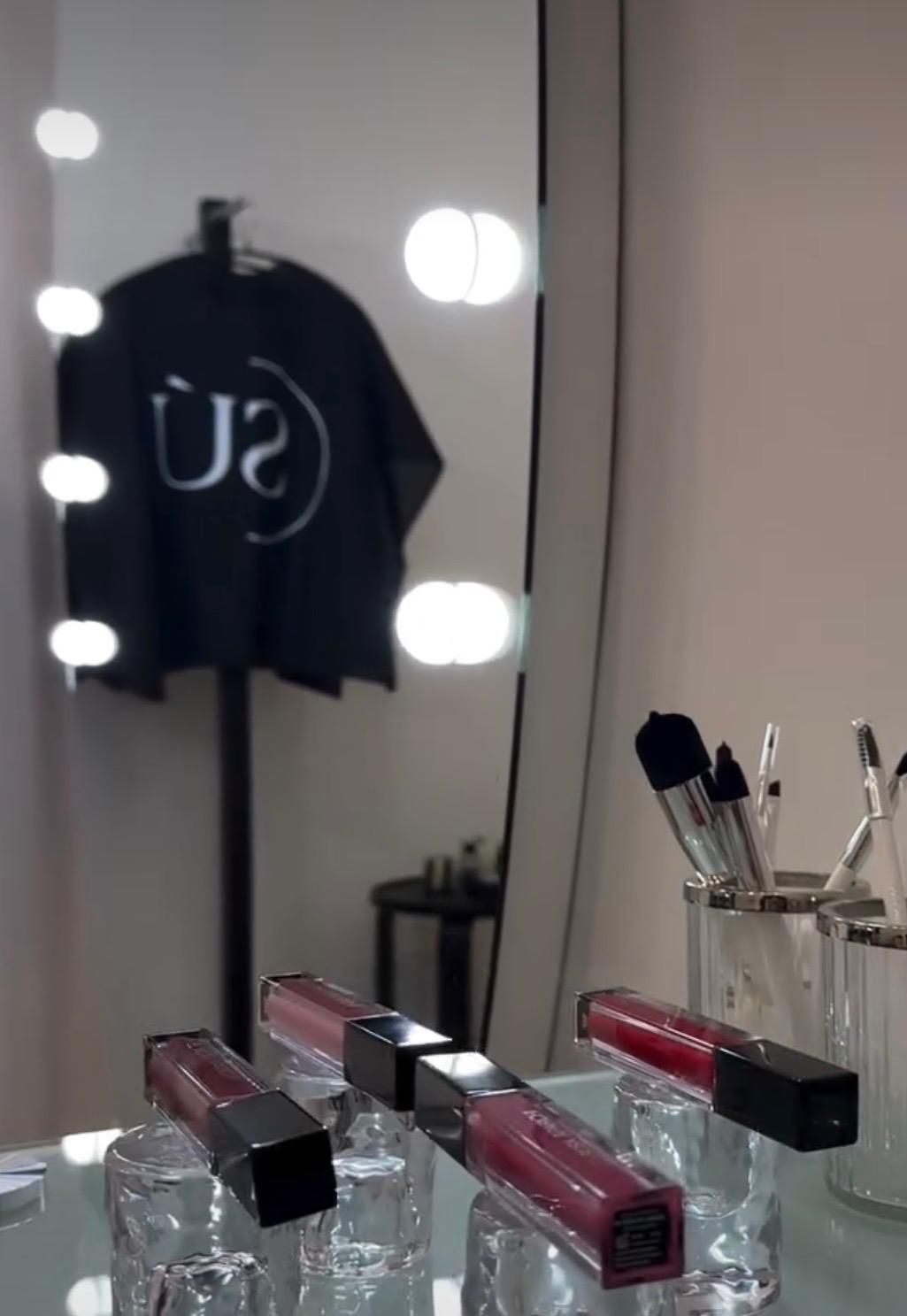 Tocador del estudio con pinceles y labiales sobre una mesa de cristal, y el reflejo de un espejo con bombillas y una capa negra con el logotipo de Sumakeup